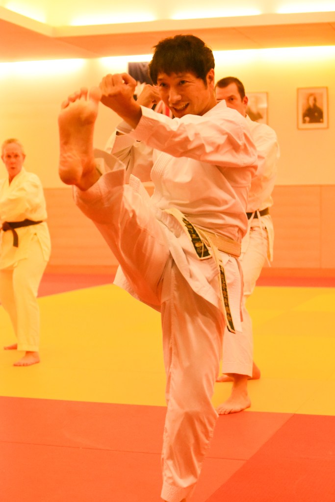 A Japanese man does a front kick and punch towards the camera while smiling. He is wearing a karate uniform and is inside a large martial arts dojo.