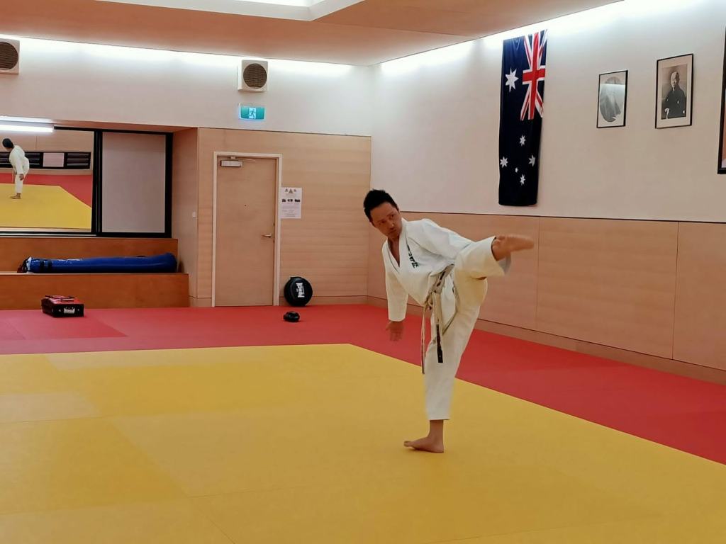 A Japanese man does a side kick in a karate uniform, inside a martial arts dojo