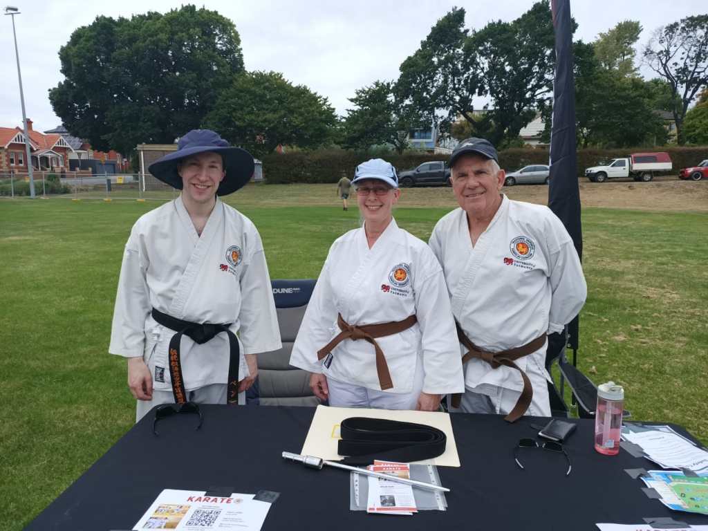 A young man, an older woman and an older man all smile for they camera. They are wearing karate uniforms and caps. On the table in front of them are several flyers and some information about TSKF. 
