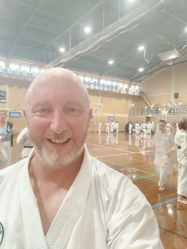 A selfie by Sensei Adrian. He is smiling after finishing 1,000 punches. In the background is a large basketball gym with many people in karate uniforms. 