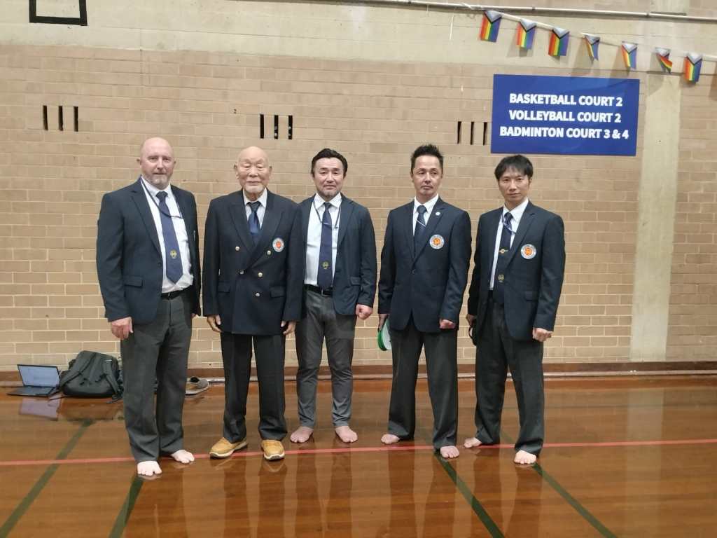 Five men, one Australian and four Japanese, wearing suits face the camera with pleasant looks. Four are not wearing shoes. They are on a basketball court. 