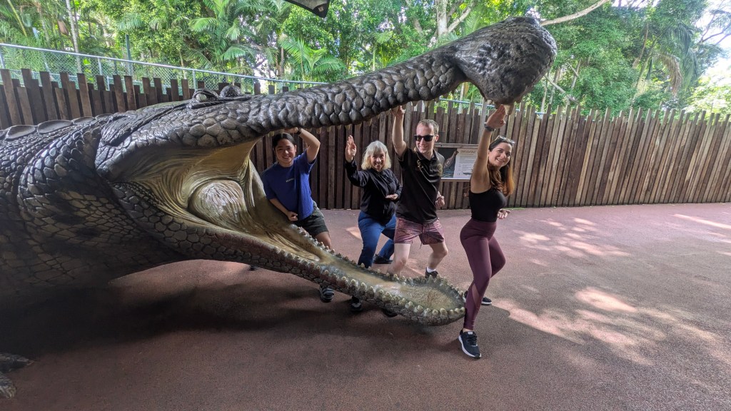 Three women and a man pretend to attack a giant prehistoric crocodile statue in karate poses. 