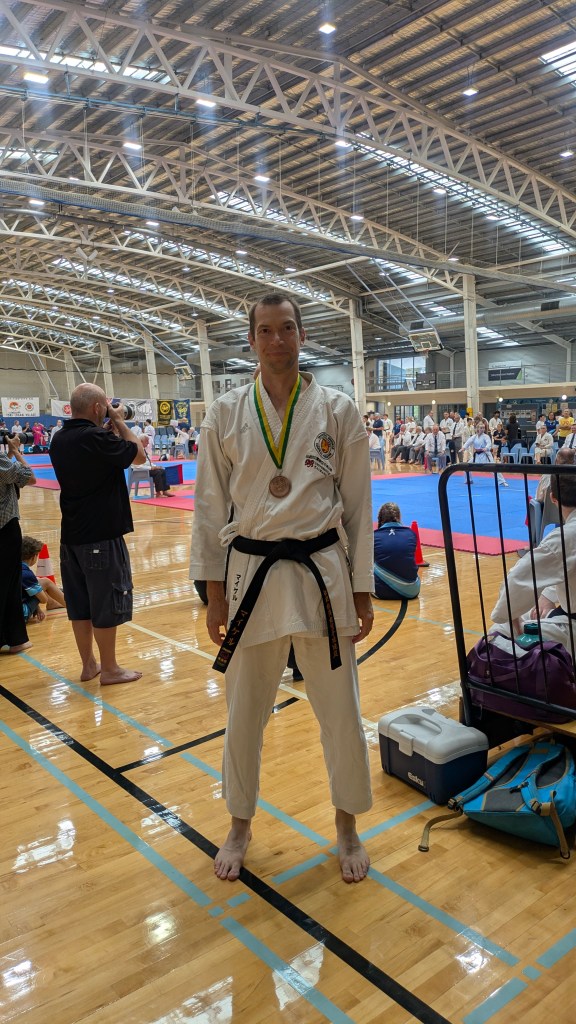 A tall man in a karate uniform smiles and is wearing a bronze medal. 